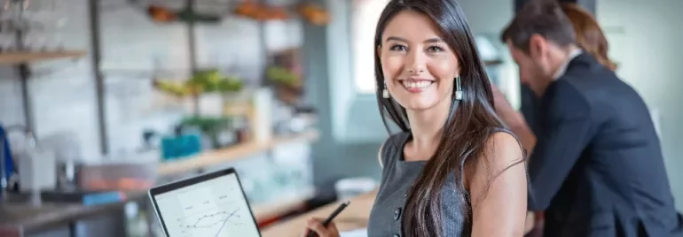 Person smiling at camera while holding a pen, with a laptop displaying graphs in an office setting.